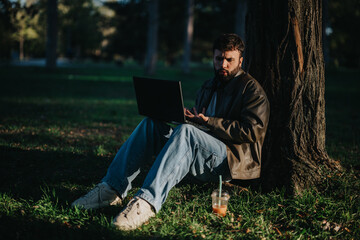 A man sits against a tree in a sunny park, focused on his laptop. He is dressed casually, enjoying...