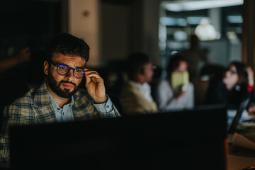 A focused businessman in glasses working late at night with colleagues in a dimly lit office...