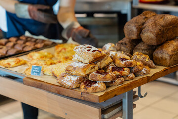 Bakery counter with assorted fresh bread, pastries, and buns on display, baker arranging baked...