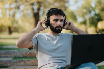 Man sitting on bench in the park, listening to music on headphones while working remotely on his...