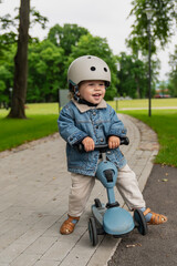 Smiling toddler wearing helmet standing on small blue scooter in park, child enjoying outdoor...
