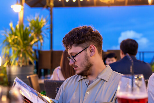 Young man with curly hair wearing glasses is focused on reading menu at outdoor restaurant, surrounded by plants and soft lighting, creating relaxed evening atmosphere. Restaurant terrace at sunset