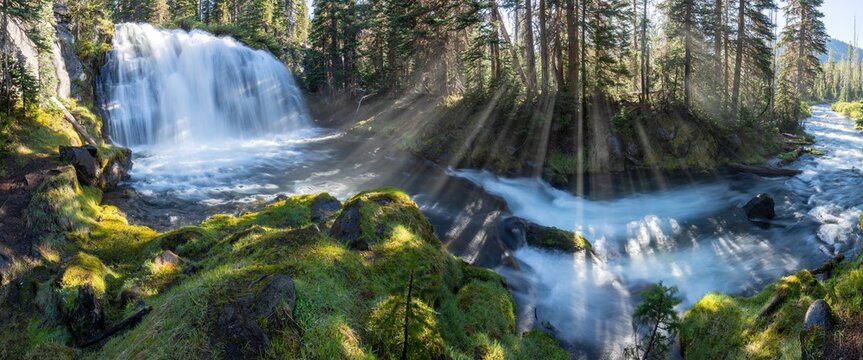 Wild mountain river with a waterfall in the canyon. The sun shines through the trees into the mist and water splinters, creating beautiful rays. A summer morning with a blue sky.