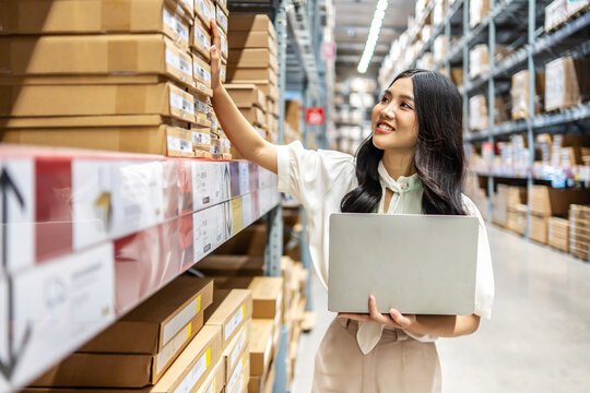Engaging in meticulous inventory management, a young asian woman professional examines products while using a laptop computer, surrounded by organized shelves filled with boxes