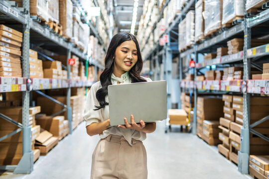 Engaging in meticulous inventory management, a young asian woman professional examines products while using a laptop computer, surrounded by organized shelves filled with boxes