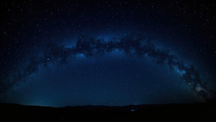Night sky panorama with the Milky Way arching across a dark blue expanse filled with countless stars above a silhouetted landscape