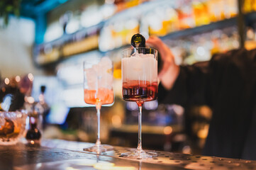Bartender pouring a cocktail into a tall glass with ice cubes on a vibrant bar counter with bottles and glassware, creating a lively nightlife ambiance.