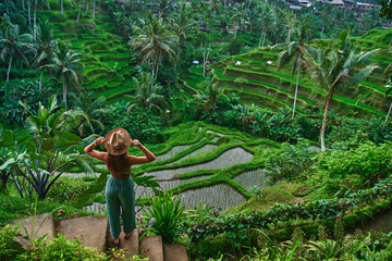 Happy carefree woman traveler wearing hat walking through picturesque green rice terraces in Ubud, Bali island, exploring and traveling to beautiful destinations