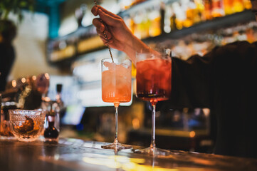 Bartender stirring two iced cocktails in tall glasses at a bar, with warm lighting and a blurred...