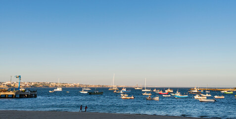 Scenic view of Cascais beach in Portugal during golden hour, with numerous colorful fishing boats and sailboats anchored in the calm Atlantic waters.