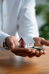 Man holding small model house and coins on wooden table representing real estate investment
