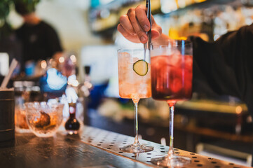 Bartender stirs cocktails at stylish bar counter, one with cucumber garnish, one red drink....
