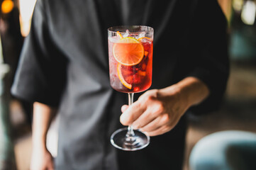 Close-up of a person in a black shirt holding a glass of red cocktail with orange slices and ice, showcasing a refreshing summer drink in stylish presentation.