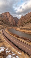 High-angle view of a winding road through a canyon, alongside a river.  Russet-colored rock formations dominate the landscape.  A light dusting of snow is visible in the foreground.  Overcast sky