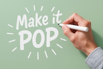 Hand adding vibrant white text "Make it POP" with a marker on a pastel green surface, illustrating creativity and visual impact.
