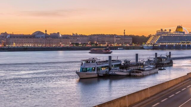 Ship and boats docked along the Garonne River in Bordeaux, France, near the ferry port. Day to night transition timelapse with waterfront reflections, traffic on the street and historic architecture