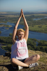A young girl sits cross-legged on a rocky outcrop, smiling as she practices yoga outdoors. The sun shines brightly over a stunning landscape filled with rivers and greenery.