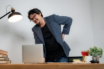Asian male office employee with back pain from prolonged sitting, showing work-related health...