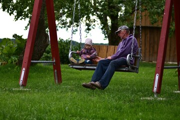 Elderly man sitting on a garden swing with a toddler girl in rural backyard. Peaceful family moment surrounded by green nature and flowers.