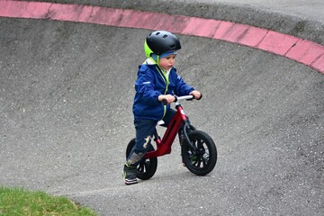 Obraz premium Active young boy in blue jacket and helmet riding a red balance bike on an outdoor pump track. Child practicing cycling skills in green park on summer day. Concept of active childhood and sport.