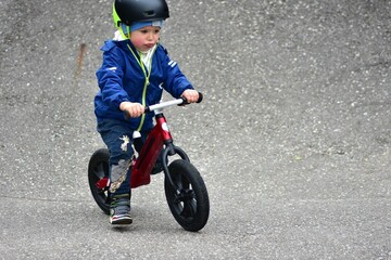 Obraz premium Active young boy in blue jacket and helmet riding a red balance bike on an outdoor pump track. Child practicing cycling skills in green park on summer day. Concept of active childhood and sport.