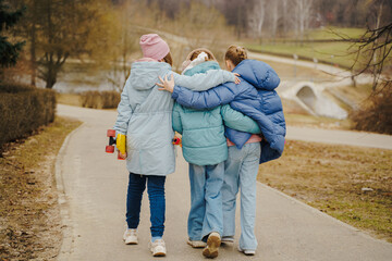 Teen girls walk together, hugging shoulders, each holding a skateboard. Jackets, jeans, sneakers...
