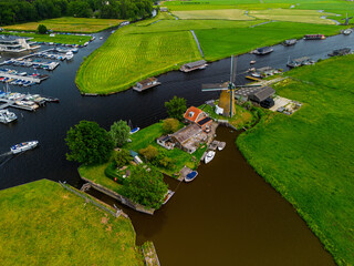 A picturesque aerial view of a Dutch windmill beside a canal with boats, surrounded by lush green fields, traditional houses, and distant waterways under a cloudy sky.