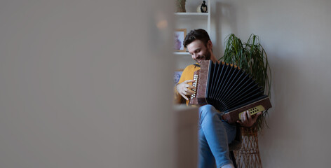 Man in a yellow sweater playing accordion while sitting at home, cozy interior with plant and shelf...
