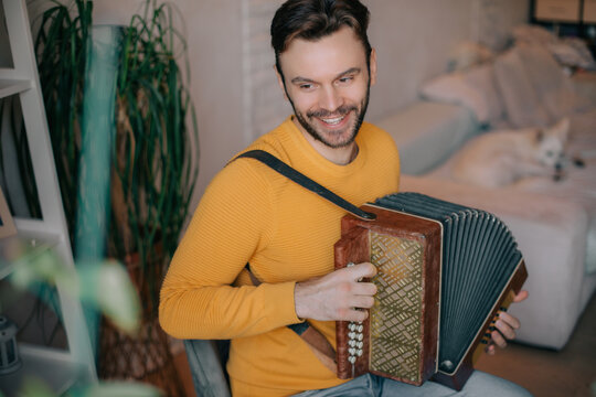 Smiling man in a yellow sweater playing accordion at home, cozy interior with plants and a white dog resting on the couch, enjoying music, folk lifestyle, and creativity.