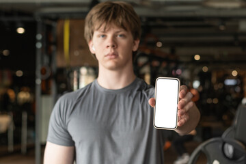 Athlete standing and displaying smartphone with white blank screen in gym setting. Mock up. Space...