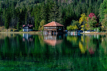 Obraz premium Charming lakeside retreat at Lago di Dobbiaco, Italy. A beautiful wooden building nestled by vibrant forests, creating stunning reflections on the clear green water. Peaceful setting.