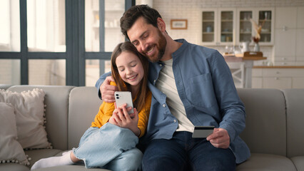 Joyful family little Caucasian girl daughter choose goods online shop with mobile phone dad man parent hold credit card clients kid father use bank app for successful payment male hug child smile home