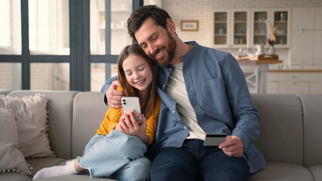 Joyful family little Caucasian girl daughter choose goods online shop with mobile phone dad man parent hold credit card clients kid father use bank app for successful payment male hug child smile home