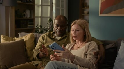Positive multiethnic senior married couple video calling friends while sitting on couch in living room, Caucasian woman holding smartphone in front of them, Black man with arm around her shoulder - Powered by Adobe