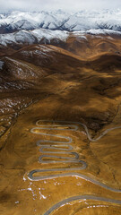 Aerial view of the winding road leading to Mount Everest from the Tibetan side, surrounded by rugged brown mountains and patches of snow under dramatic skies.