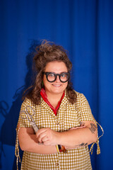 Fashion designer holding scissors, arms crossed, smiling confidently while wearing glasses against blue backdrop