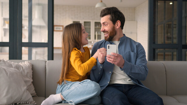 Happy positive Caucasian family daughter dad using mobile phone man guy winning child little smiling girl celebrating at home male parent typing screen victory in online game father give kid high five