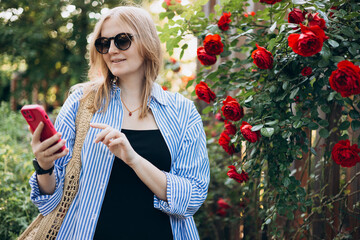 A portrait of a smiling beautiful 30s woman texting with her phone. Summer time. Young female in casual clothing standing outdoor