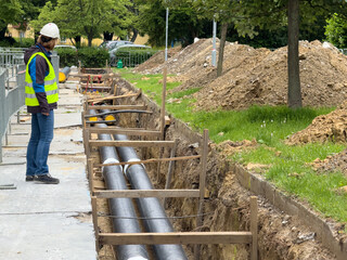 Female engineer in protective gear inspects district heating pipeline installation in urban green zone. Ideal for: energy infrastructure, female engineers, civil works documentation, editorial use.