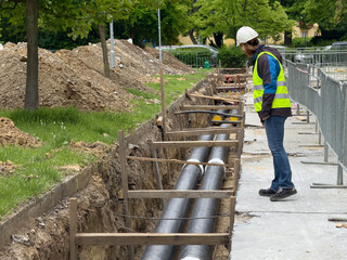 Female engineer in protective gear inspects district heating pipeline installation in urban green zone. Ideal for: energy infrastructure, female engineers, civil works documentation, editorial use.