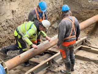 Three utility workers installing a pre-insulated steel pipe at a district heating construction site. Precise alignment and teamwork during underground pipe assembly for thermal energy distribution.
