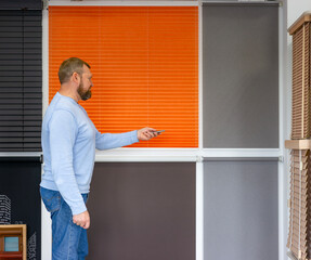 Man in a sunshades company office uses a remote control to operate automated blinds. He stands beside a wall, displaying featuring a variety of blinds and curtains types, materials, and color options.