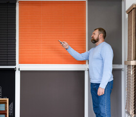 In a sunshade systems showroom, a man demonstrates remote-controlled blinds. Bright orange pleated blinds and gray roller shades are part of a smart, stylish, and functional window treatment display.