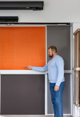 Man in a light blue sweater demonstrates motorized blinds using a remote control in a modern showroom. Display features pleated, roller, and wooden blinds designed for smart light and privacy control.