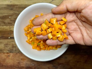 photo of a woman's palm showing at close range carrot slices cut into small squares in a white plastic bowl with a dark brown wooden table background, carrot slices ready to be cooked or pickled