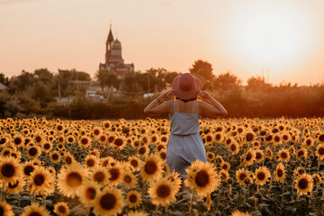 Beautiful happy young girl in blue dress and hat in sunflower field at sunset