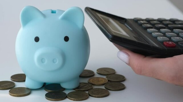 Hand counting on calculator next to piggy bank and coins on white table. Saving concept