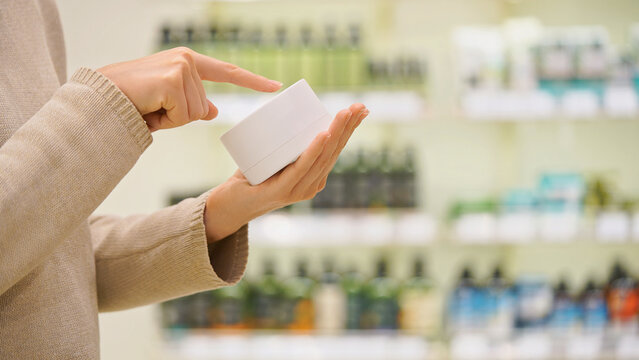 Woman holding a cosmetic jar in a beauty store, reading the label and examining ingredients. Moisturizing anti-aging face cream or nourishing hair mask. Concept of conscious choice, skincare, haircare