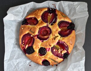 A round cake topped with slices of plums and blackberries is placed on a piece of white parchment...