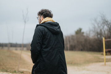 A young man with glasses and a coat stares across an open field on a cloudy day. The outdoor...